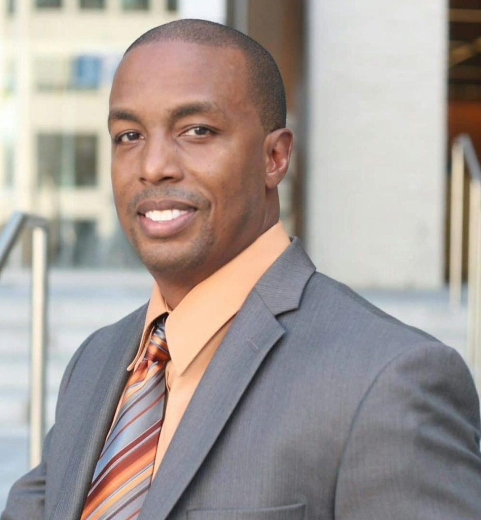John Smith in a suit and tie poses outdoors in a modern urban environment.